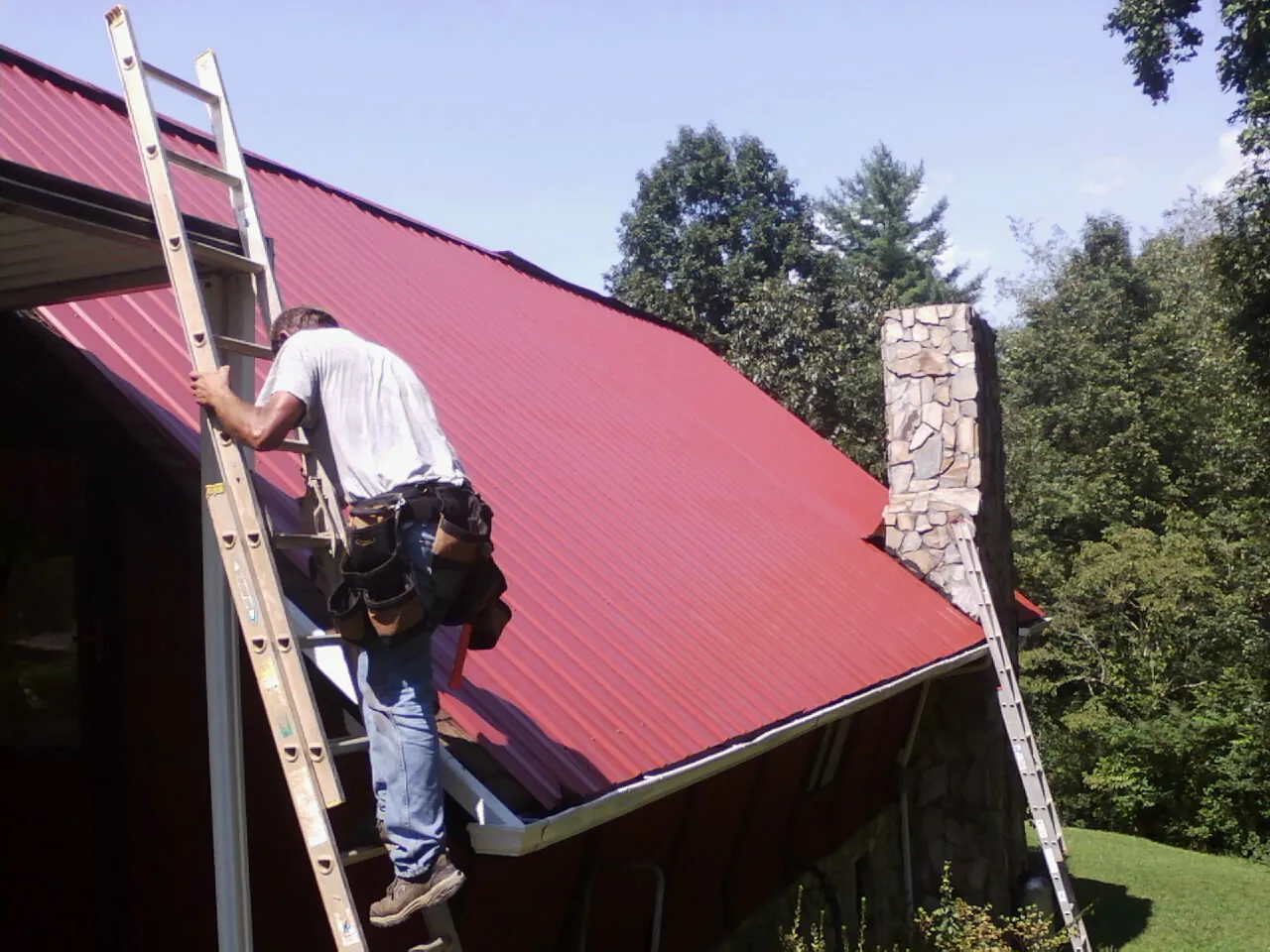 Roofer on a ladder beside a red metal roof and stone chimney — Soffit & Fascia Repair by Roofing Brothers in Ridgecrest